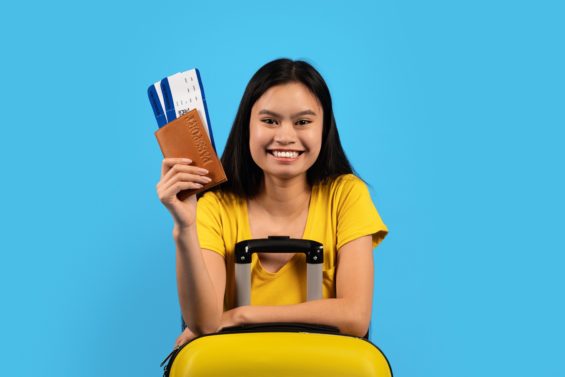 Portrait of smiling young korean lady with down syndrome in yellow t-shirt with suitcase show passport, air ticket